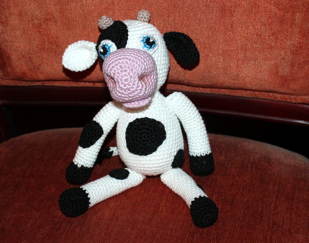 Detailansicht einer sitzenden Amigurumi-Kuh mit schwarzen Flecken, rosa Schnauze und blauen Augen