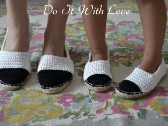 Zwei Paar gehäkelte Espadrilles in Weiß mit schwarzer Zehenkappe und Jutesohle auf floralem Teppich; ein Paar für Erwachsene und ein Paar für Kinder.