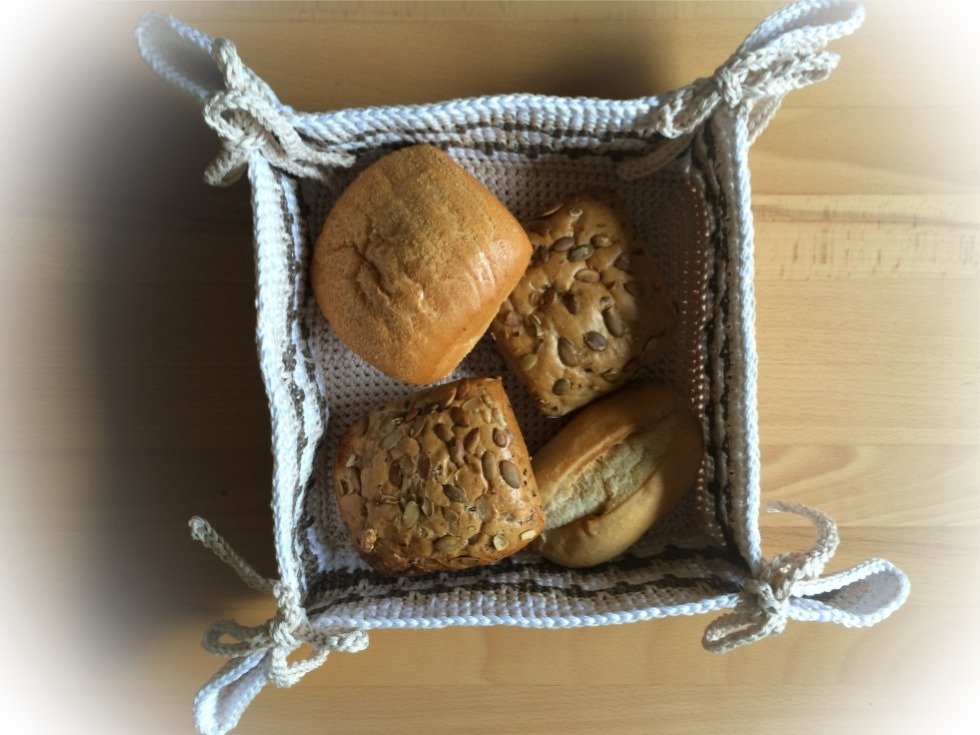 Gehäkelter Korb mit Schleifen, von oben gezeigt, gefüllt mit vier Brötchen auf einem Holztisch.