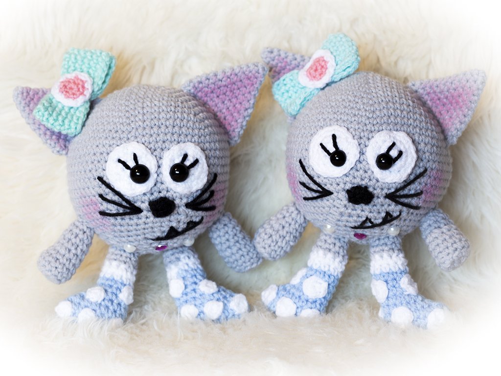 Zwei gehäkelte graue Amigurumi-Katzen mit türkisfarbenen Schleifen und gepunkteten Beinen auf hellem Hintergrund.