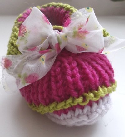 Gestrickter Babyschuh in Pink mit grünem Rand, weißer Sohle und geblümter Stoffschleife.