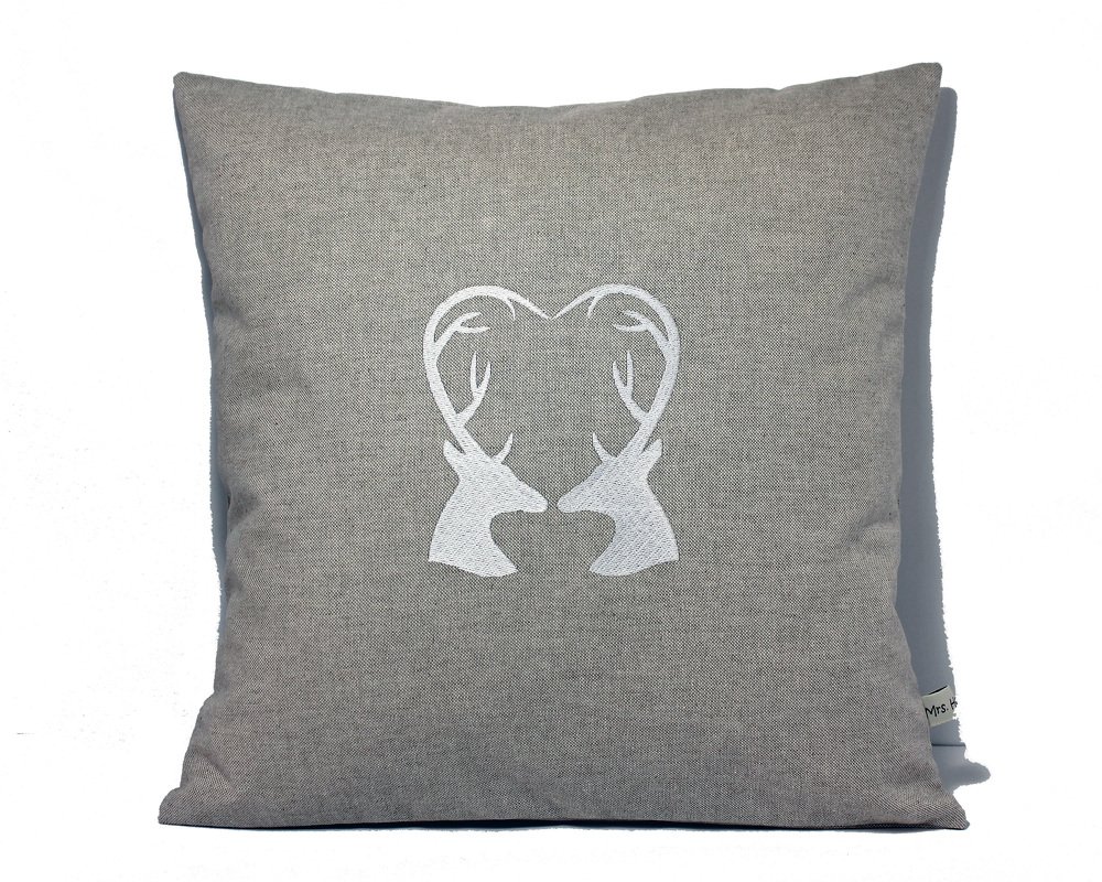 Graues Kissen mit weißer Stickerei: zwei Hirschköpfe, deren Geweihe ein Herz bilden.