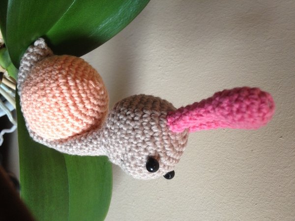 Gehäkelte Amigurumi-Schnecke mit beigem Körper, pfirsichfarbenem Haus und pinkem Fühler auf grünem Blatt.