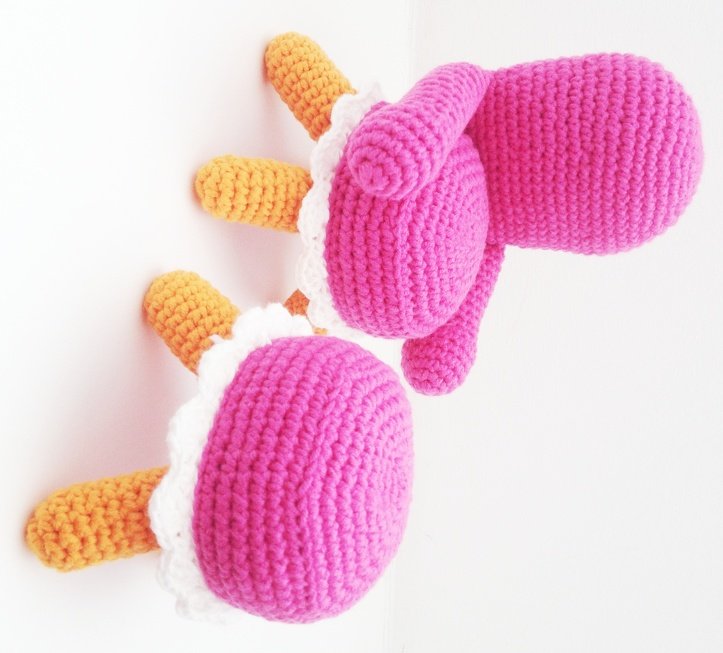 Zwei rosa gehäkelte Amigurumi mit orangefarbenen Beinchen und weißer Zierborte
