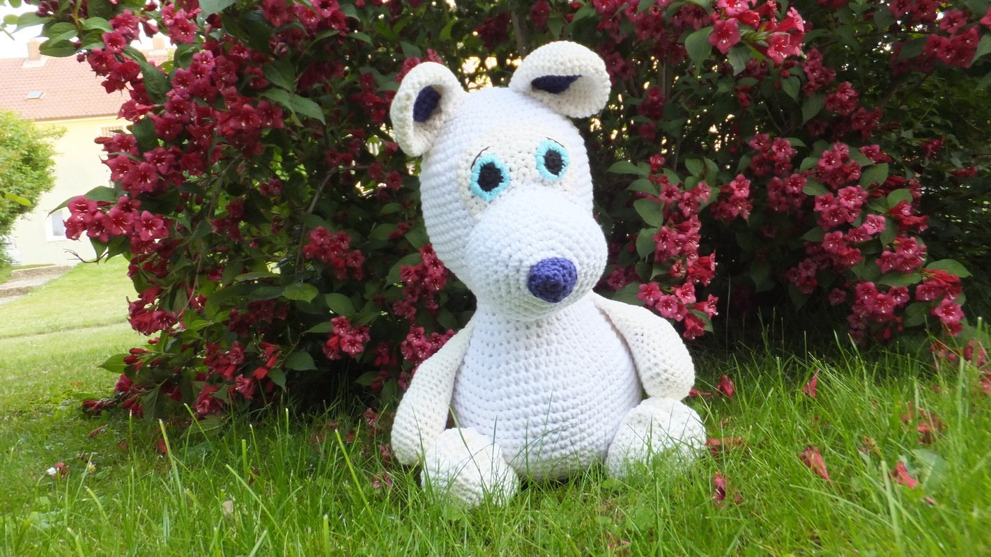 Weißer gehäkelter Amigurumi-Bär mit hellblauen Augen und lila Nase im grünen Gras vor einem Busch mit roten Blüten.