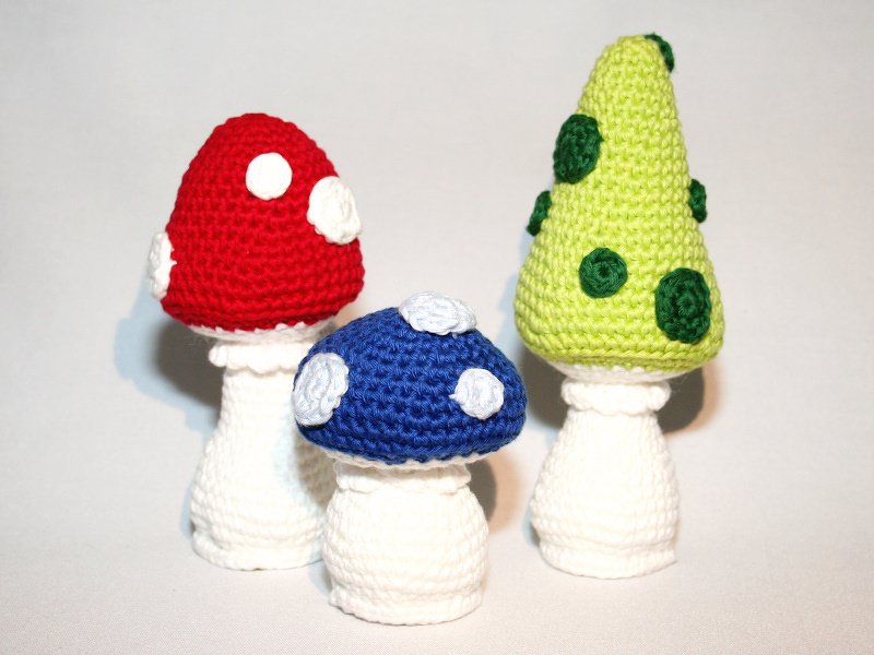 Three crocheted mushrooms: red and blue rounded spotted caps and a lime-green cone cap with dark green spots, all on white stems
