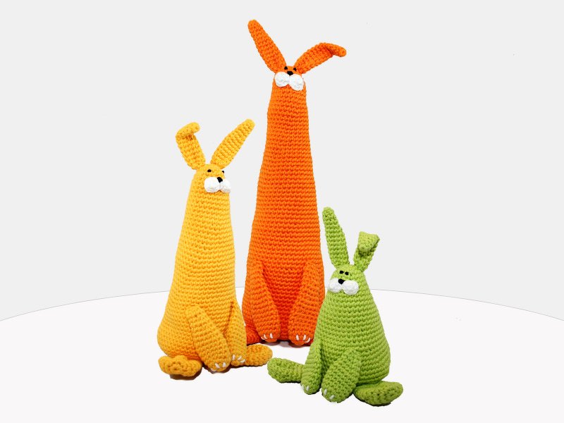 Three crocheted amigurumi rabbits in orange, yellow and green with long ears on a white surface
