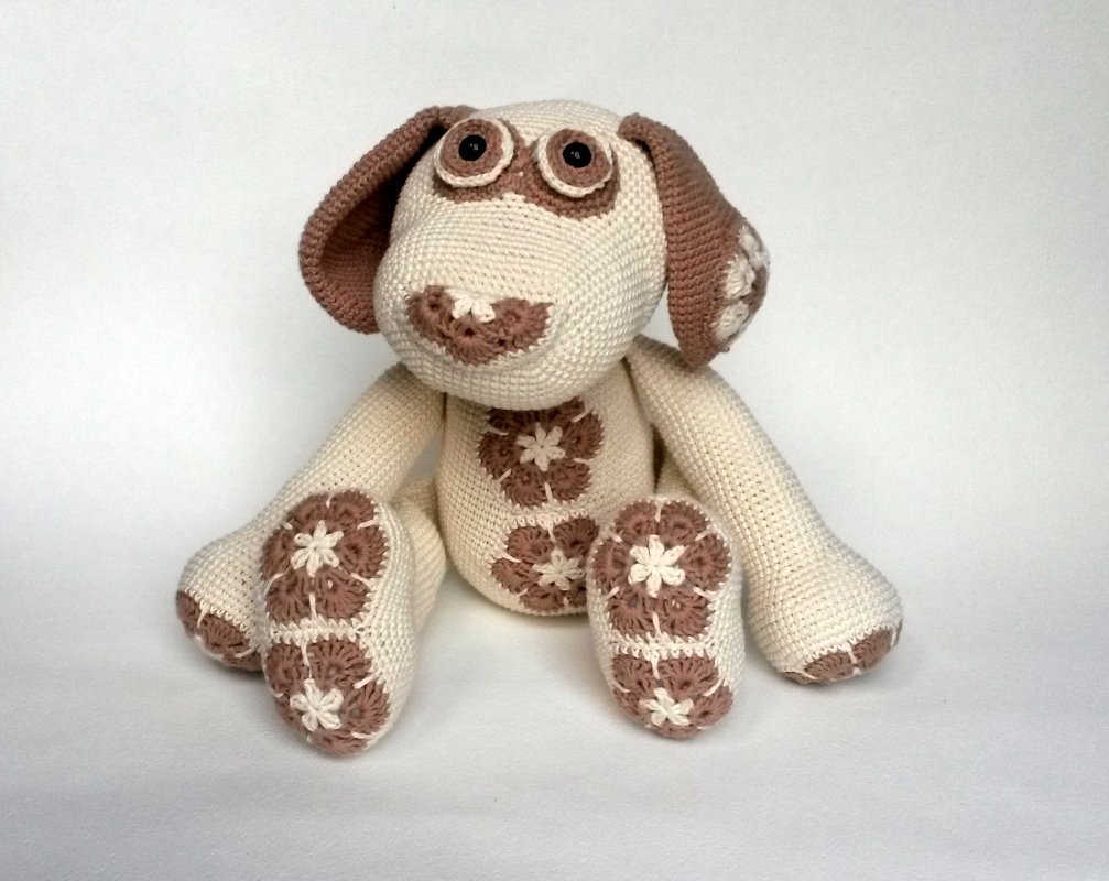 Sitzender Amigurumi-Hund in Creme und Braun mit blütenförmigen Häkel-Applikationen auf Bauch und Pfoten