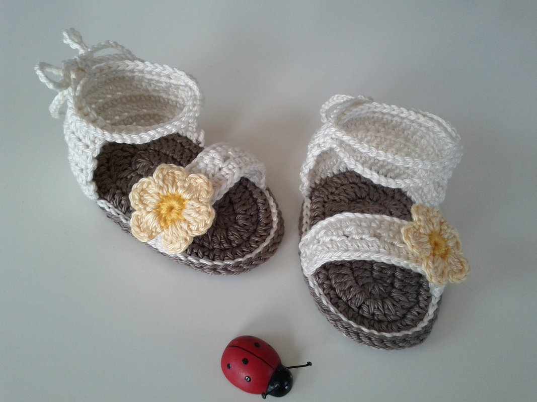 Gehäkelte Babysandalen in Creme und Braun mit gelber Blüte und Schnürband, daneben kleines Marienkäfer-Spielzeug
