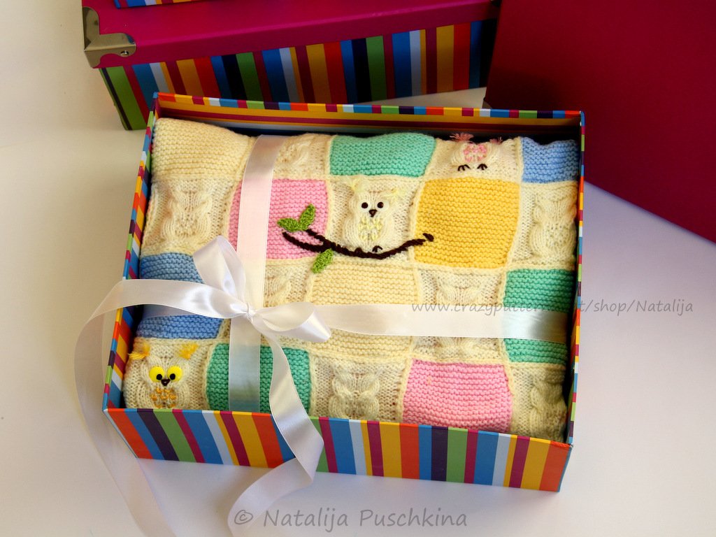Gefaltete gestrickte Patchworkdecke mit Eulenmotiven in einer bunten Geschenkbox mit weißer Satinschleife.