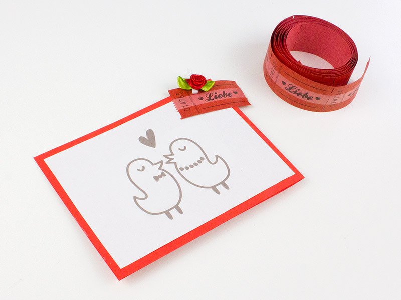 Selbstgemachte weiße Karte mit rotem Rand, zwei gezeichnete Vögel, Herz und rotes Zierband mit 'Liebe'