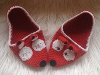 Rote gehäkelte Hausschuhe mit Fuchsapplikation auf flauschigem Untergrund.