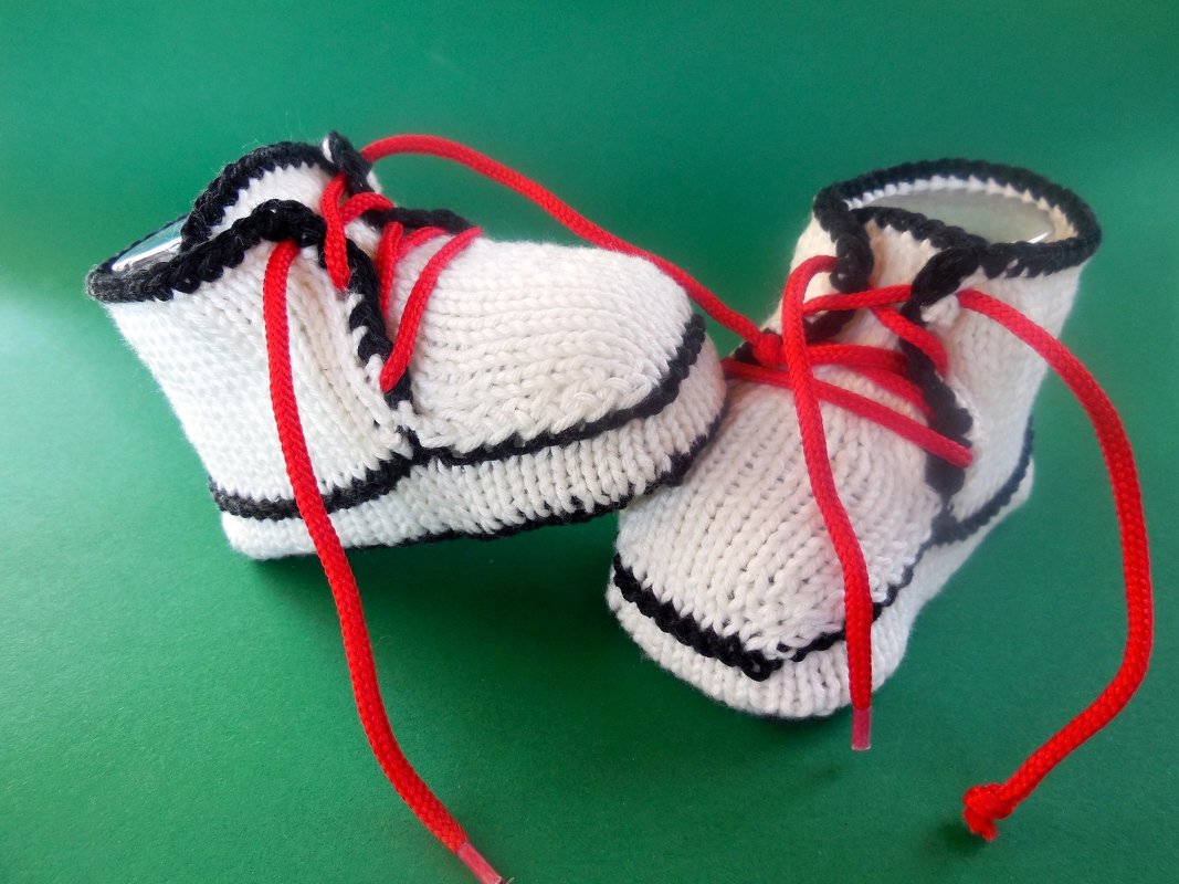 Zwei gestrickte weiße Babyschuhe mit schwarzer Einfassung und roten Schnürsenkeln auf grünem Hintergrund