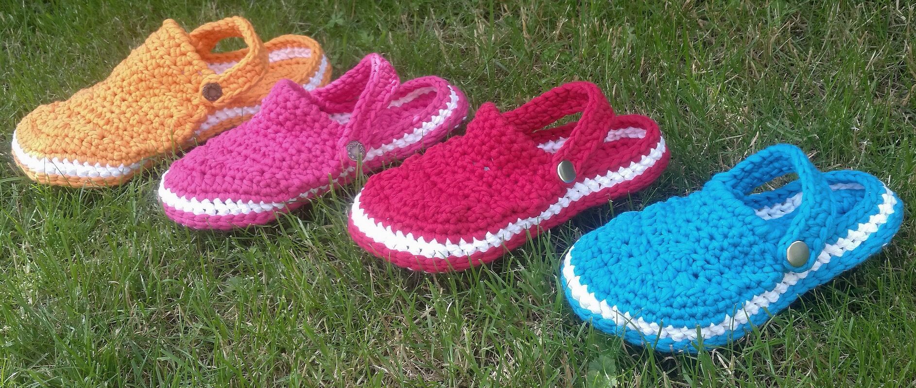 Vier gehäkelte Slipper in Orange, Pink, Rot und Blau auf Gras mit weißem Sohlensaum