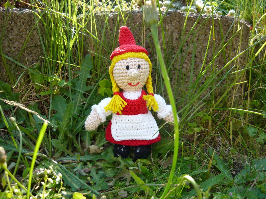 gehäkelte Amigurumi-Puppe in rotem Kleid mit gelben Zöpfen, sitzend im Gras