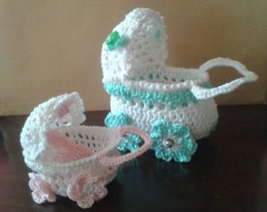 Zwei gehäkelte Mini-Kinderwagen in Weiß mit rosa und mintfarbenen Verzierungen und Blumenapplikationen