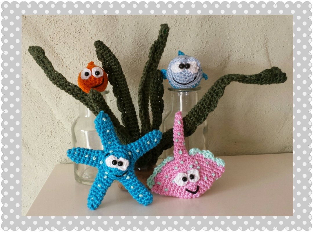 Gehäkelte Amigurumi-Meerestiere: blauer Seestern, rosa Rochen, zwei kleine Fische und grünes Seegras vor Glasvasen