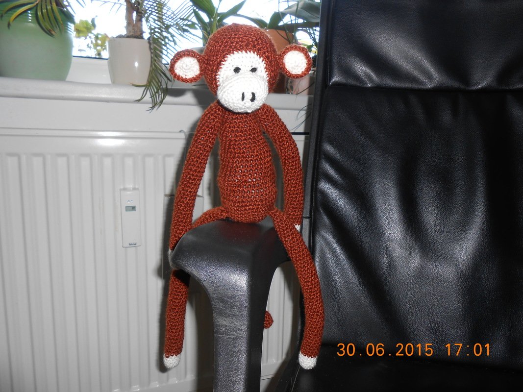 Brauner gehäkelter Amigurumi-Affe mit weißer Schnauze und sehr langen Armen sitzend auf schwarzer Stuhlarmlehne