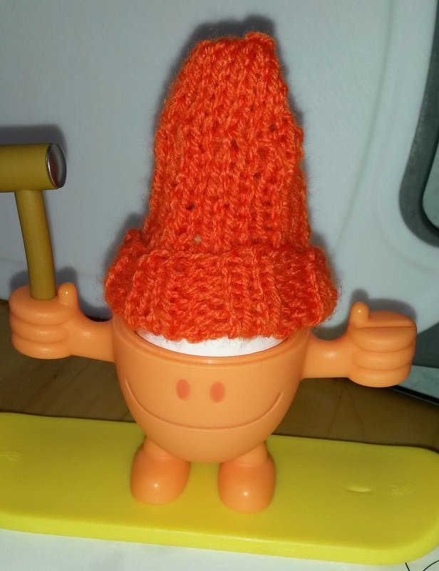 Orange gestrickter Eierwärmer auf einem orangefarbenen Eierbecher mit Gesicht auf gelber Unterlage