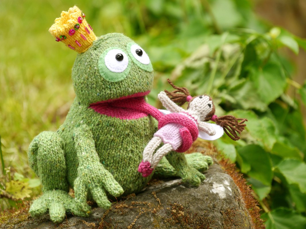 Hand-knit green frog amigurumi wearing a yellow beaded crown holding a small pink-and-white fairy doll on a mossy rock with green foliage behind.