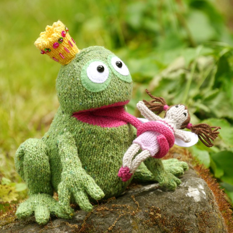 Handgestrickter Froschkönig mit Krone hält eine kleine gestrickte Fee an seiner Zunge.