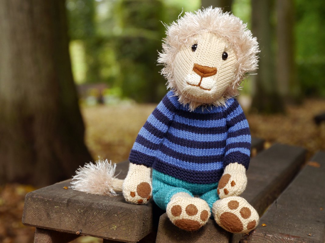 Knitted amigurumi lion toy with a fluffy mane, wearing a blue striped sweater and teal pants, seated on a wooden bench.
