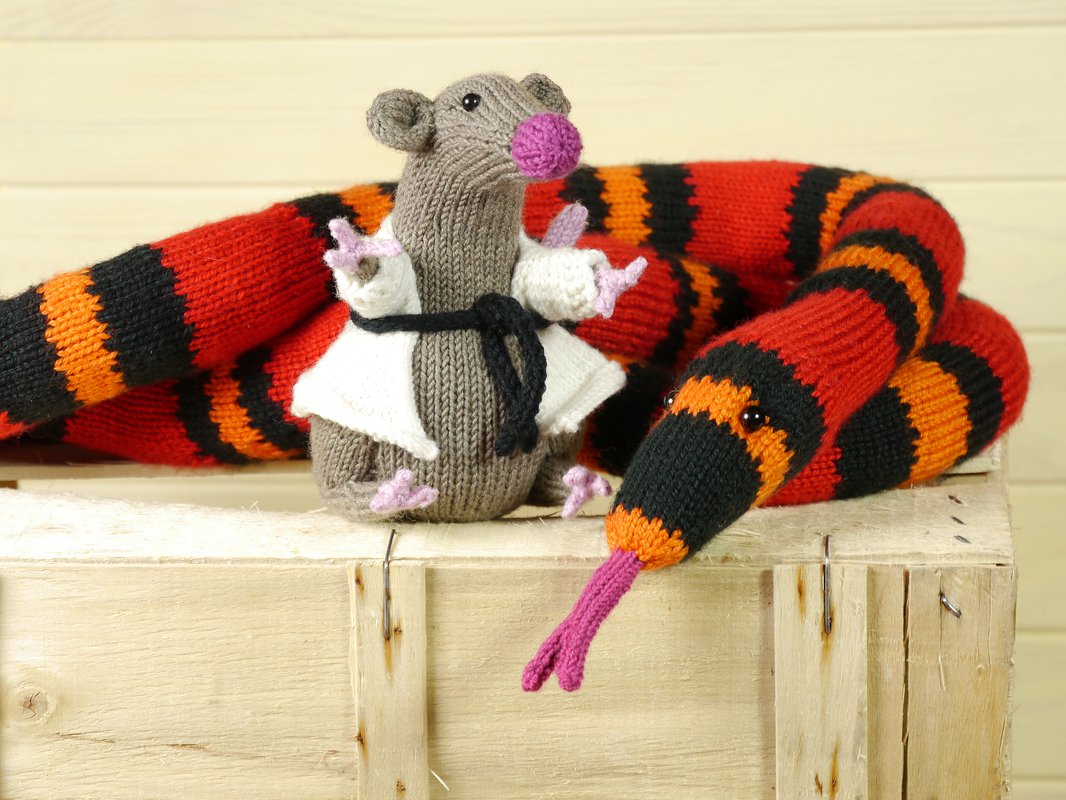 Gestrickte graue Maus im weißen Kimono mit schwarzem Gürtel neben rot-orange-schwarz gestreifter Schlange mit rosa Zunge auf Holzkiste.