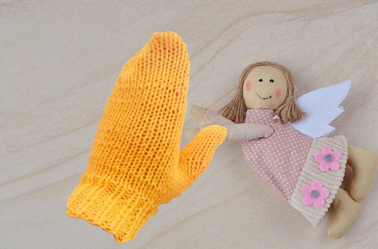 Gelber gestrickter Kinderfäustling neben einer Stoffpuppe mit rosa Kleid und weißen Flügeln auf hellem Hintergrund.