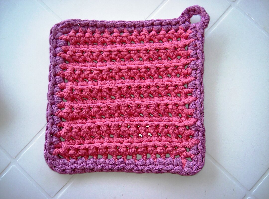 Quadratischer handgehäkelter Topflappen in Rosa und Lila mit Aufhängeschlaufe auf weißer Fliese