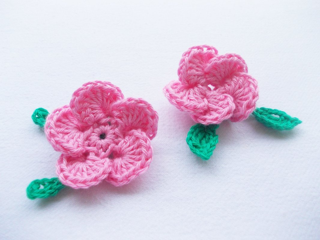 zwei rosa gehäkelte Blumenapplikationen mit grünen Blättern auf weißem Hintergrund