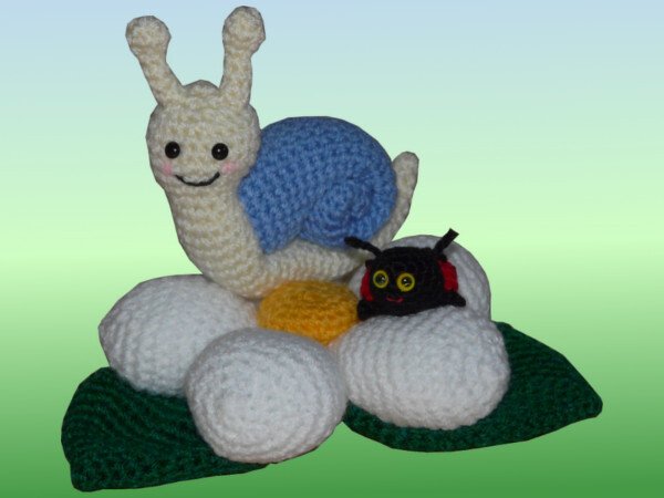Gehäkelte Amigurumi-Schnecke mit blauem Haus und kleine schwarze Raupe auf weißer gehäkelter Blume