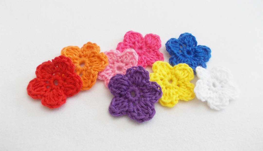 Acht gehäkelte Blumen in Regenbogenfarben auf weißem Hintergrund.