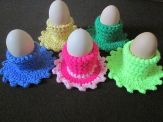 Fünf gehäkelte Eierwärmer in leuchtenden Farben mit je einem Ei auf dunklem Hintergrund