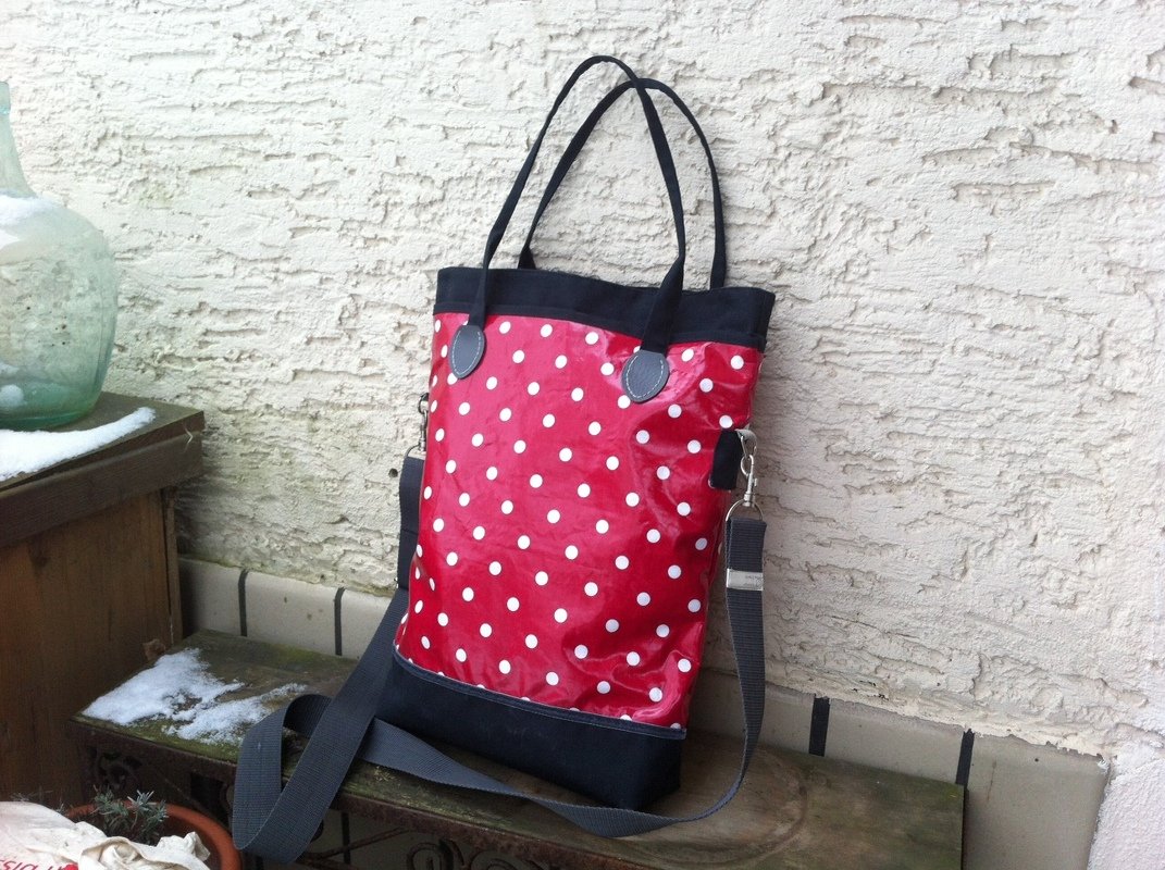 Glänzende rote Einkaufstasche mit weißen Punkten, schwarzem Rand und Boden, Tragegriffen und abnehmbarem Schultergurt, an einer strukturierten Wand gelehnt.