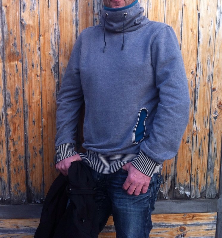 Mann in grauem Pullover mit hohem Kragen, blauer Innentasche und gestreiften Bündchen vor Holzwand