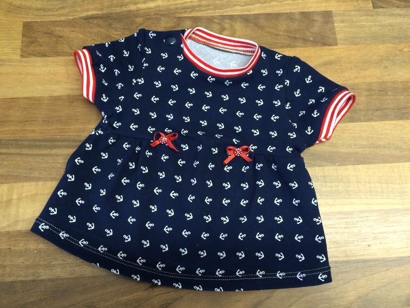 Marineblaues Babykleid mit weißem Anker-Print, rot-weiß gestreiften Bündchen und roten Schleifen.