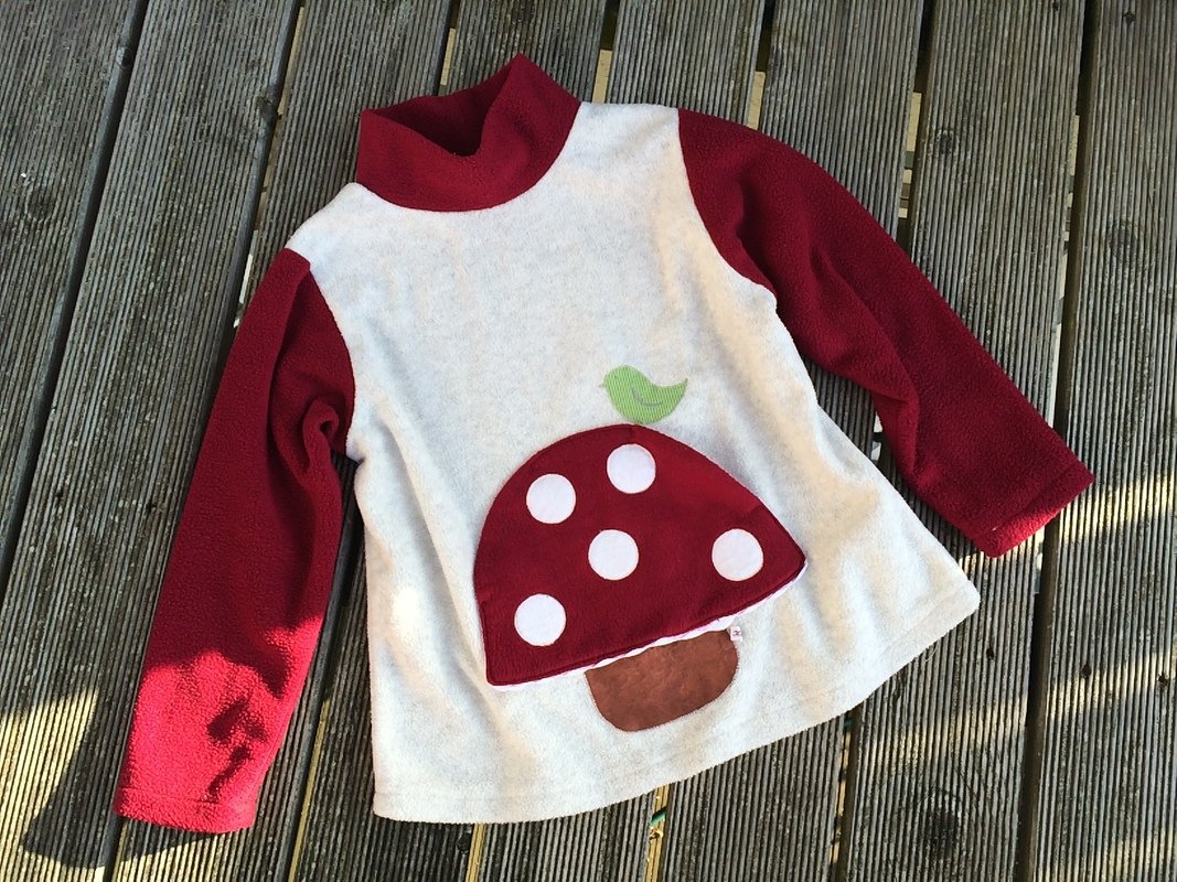 Nahaufnahme eines genähten Kinderpullovers mit roter Fliegenpilz-Applikation, beiger Vorderseite und roten Ärmeln auf Holzdielen