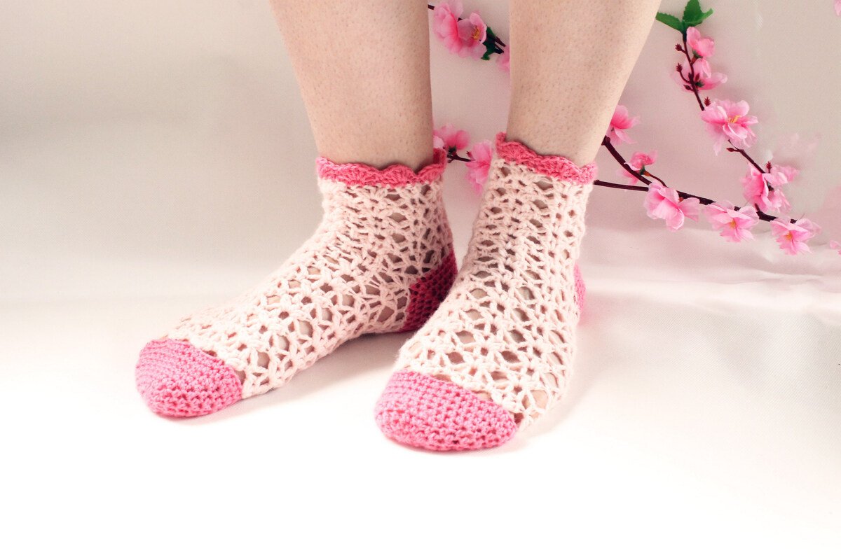 Kurze, luftige gehäkelte Socken in hellrosa mit pinker Ferse und Zehe sowie gezacktem Bündchen, auf hellem Hintergrund mit rosa Blütenzweig