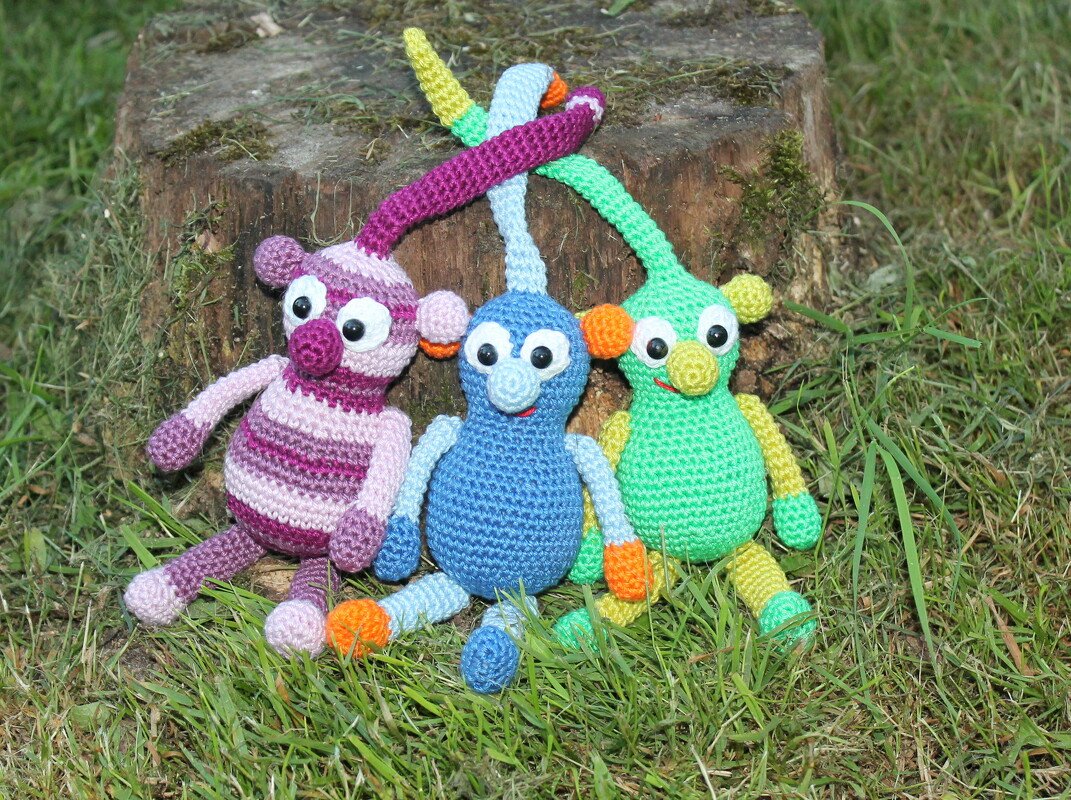Drei bunte gehäkelte Figuren mit langen Schlaufen auf Gras vor einem Baumstumpf