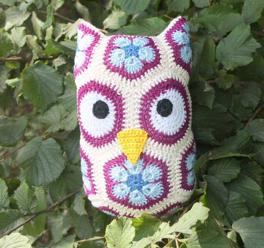 Gehäkeltes Eulen-Kissen aus Granny- und African-Flower-Motiven, weiße Grundfarbe mit magenta- und hellblauen Akzenten, gelber Schnabel, schwarze Augen vor grünen Blättern