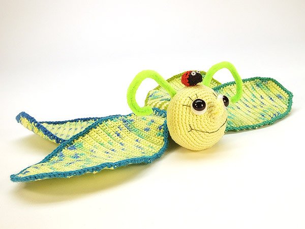 Gehäkelte Libelle in Gelb und Grün mit blau akzentuierten Flügeln und kleinem Marienkäfer auf hellem Hintergrund