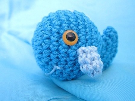Gehäkelter blauer Amigurumi-Wal mit gelbem Auge und hellblauer Flosse auf blauem Stoff.
