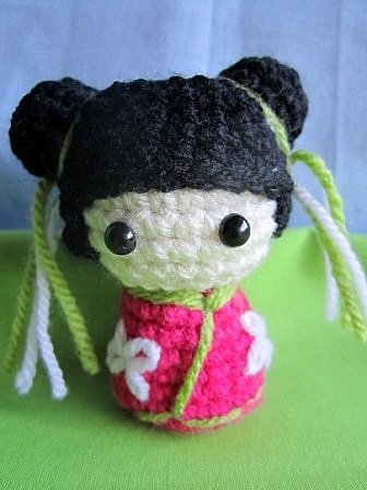 Handgehäkelte kleine Amigurumi-Puppe mit dunklen Dutts, großen schwarzen Augen und pinkem Kleid mit weißen Blüten.