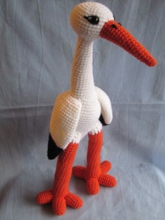 Gehäkelter weißer Storch mit langem orangefarbenem Schnabel und orangefarbenen Beinen, schwarzen Augen und dunklen Flügelspitzen vor hellem Hintergrund.