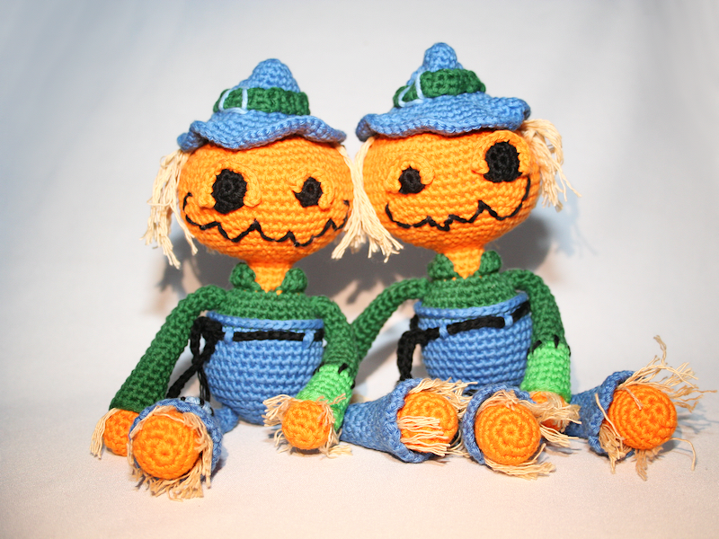 Zwei gehäkelte Kürbispuppen mit blauen Hüten, grünen Ärmeln und Garnhaaren sitzen nebeneinander