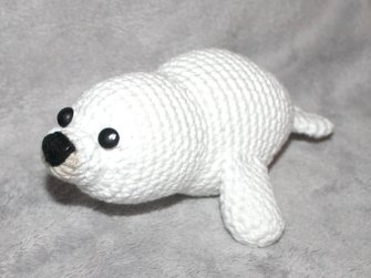 Weißes gehäkeltes Robben-Amigurumi mit schwarzen Knopfaugen und schwarzer Nase auf grauem Stoff.