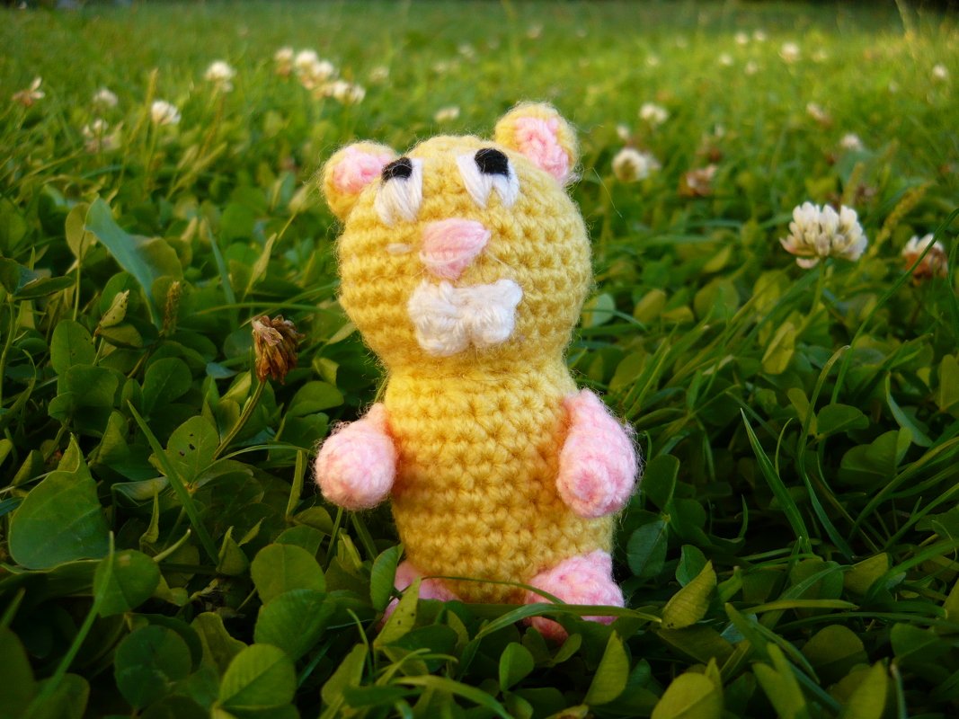 Gehäkelter gelber Hamster mit rosa Details und weißen Zähnen auf einer Klee‑Wiese.
