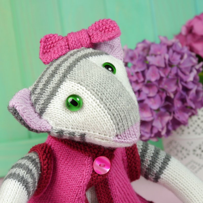Striped white and grey knitted amigurumi wearing a pink cardigan and bow, with green glass eyes.