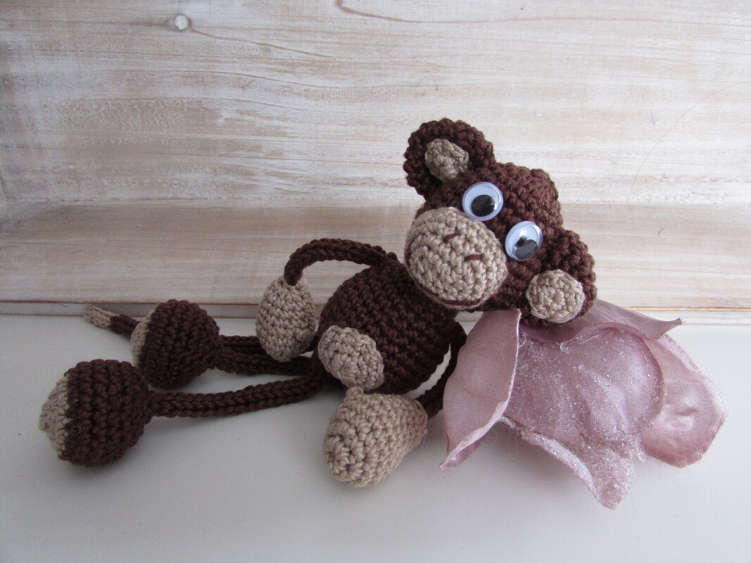 Gehäkelter brauner Amigurumi-Affe mit beigen Details, großen Kulleraugen und rosa Blütenapplikation, liegend