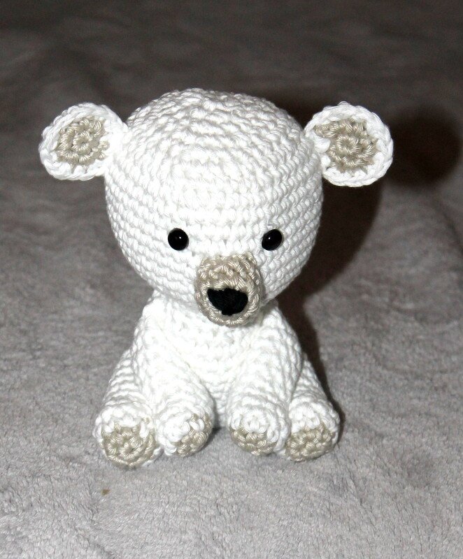 Weißes, gehäkeltes Amigurumi-Eisbärchen mit beigen Details an Ohren, Schnauze und Pfoten sowie schwarzen Augen und Nase, sitzend auf grauem Untergrund.